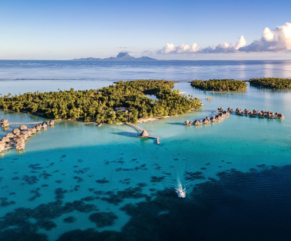 Aerial view of a lush tropical island surrounded by crystal-clear turquoise lagoon waters, with rows of overwater bungalows extending from the shoreline on curved wooden walkways. A small boat travels across the lagoon in the foreground, while distant mountainous islands and scattered clouds sit on the horizon under a bright blue sky.