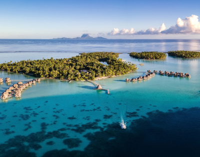 Aerial view of a lush tropical island surrounded by crystal-clear turquoise lagoon waters, with rows of overwater bungalows extending from the shoreline on curved wooden walkways. A small boat travels across the lagoon in the foreground, while distant mountainous islands and scattered clouds sit on the horizon under a bright blue sky.