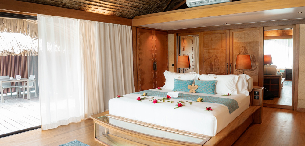 Spacious tropical resort bedroom with a large king-sized bed decorated with fresh flowers, surrounded by warm timber furnishings and carved wooden cabinetry. Floor-to-ceiling curtains open to a private outdoor deck with seating, while a glass viewing panel at the foot of the bed reveals the water below, highlighting the overwater bungalow setting.