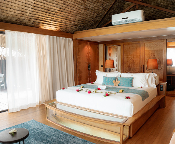 Spacious tropical resort bedroom with a large king-sized bed decorated with fresh flowers, surrounded by warm timber furnishings and carved wooden cabinetry. Floor-to-ceiling curtains open to a private outdoor deck with seating, while a glass viewing panel at the foot of the bed reveals the water below, highlighting the overwater bungalow setting.