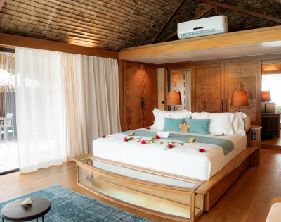 Spacious tropical resort bedroom with a large king-sized bed decorated with fresh flowers, surrounded by warm timber furnishings and carved wooden cabinetry. Floor-to-ceiling curtains open to a private outdoor deck with seating, while a glass viewing panel at the foot of the bed reveals the water below, highlighting the overwater bungalow setting.