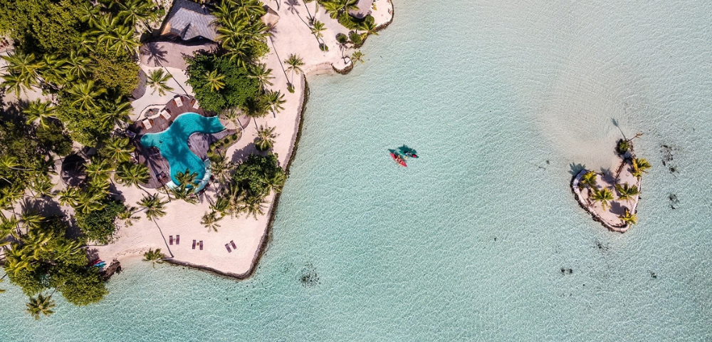 Aerial view of a tropical island fringed with white sand and dense palm trees, surrounded by a shallow turquoise lagoon with crystal-clear water. A freeform swimming pool sits near the shoreline, kayaks float in the lagoon, and a tiny palm-covered islet appears offshore to the right.