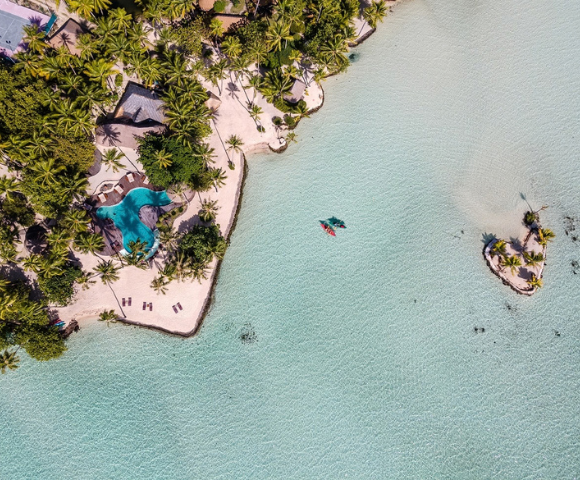 Aerial view of a tropical island fringed with white sand and dense palm trees, surrounded by a shallow turquoise lagoon with crystal-clear water. A freeform swimming pool sits near the shoreline, kayaks float in the lagoon, and a tiny palm-covered islet appears offshore to the right.