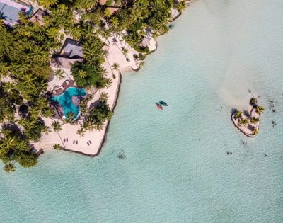 Aerial view of a tropical island fringed with white sand and dense palm trees, surrounded by a shallow turquoise lagoon with crystal-clear water. A freeform swimming pool sits near the shoreline, kayaks float in the lagoon, and a tiny palm-covered islet appears offshore to the right.