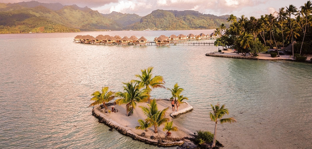 A small palm-covered sand islet sits in a calm tropical lagoon, with overwater bungalows stretching across the water in the background and lush green mountains beyond. Warm golden light reflects across the lagoon, highlighting the island resort setting and the surrounding turquoise shallows.