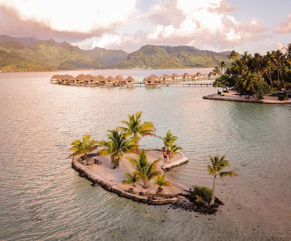 A small palm-covered sand islet sits in a calm tropical lagoon, with overwater bungalows stretching across the water in the background and lush green mountains beyond. Warm golden light reflects across the lagoon, highlighting the island resort setting and the surrounding turquoise shallows.