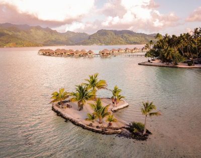 A small palm-covered sand islet sits in a calm tropical lagoon, with overwater bungalows stretching across the water in the background and lush green mountains beyond. Warm golden light reflects across the lagoon, highlighting the island resort setting and the surrounding turquoise shallows.