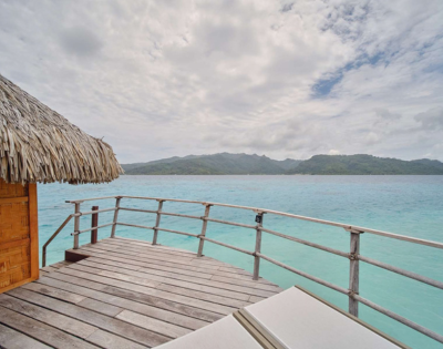 Wooden deck of an overwater bungalow with a thatched roof overlooks calm turquoise ocean water, with a lush green mountainous island stretching across the horizon. The scene is bright but slightly overcast, with textured clouds filling much of the sky and a lounge chair partially visible in the foreground.