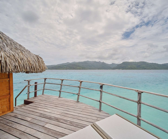 Wooden deck of an overwater bungalow with a thatched roof overlooks calm turquoise ocean water, with a lush green mountainous island stretching across the horizon. The scene is bright but slightly overcast, with textured clouds filling much of the sky and a lounge chair partially visible in the foreground.