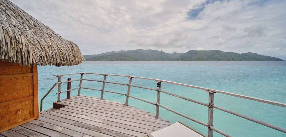 Wooden deck of an overwater bungalow with a thatched roof overlooks calm turquoise ocean water, with a lush green mountainous island stretching across the horizon. The scene is bright but slightly overcast, with textured clouds filling much of the sky and a lounge chair partially visible in the foreground.