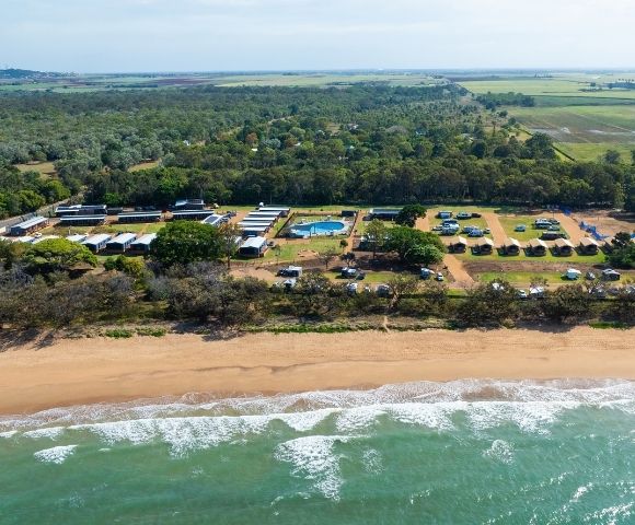 Aerial view of a beachside campground with multiple cabins, a swimming pool, and scattered tents and RVs surrounded by trees and open fields in the background.