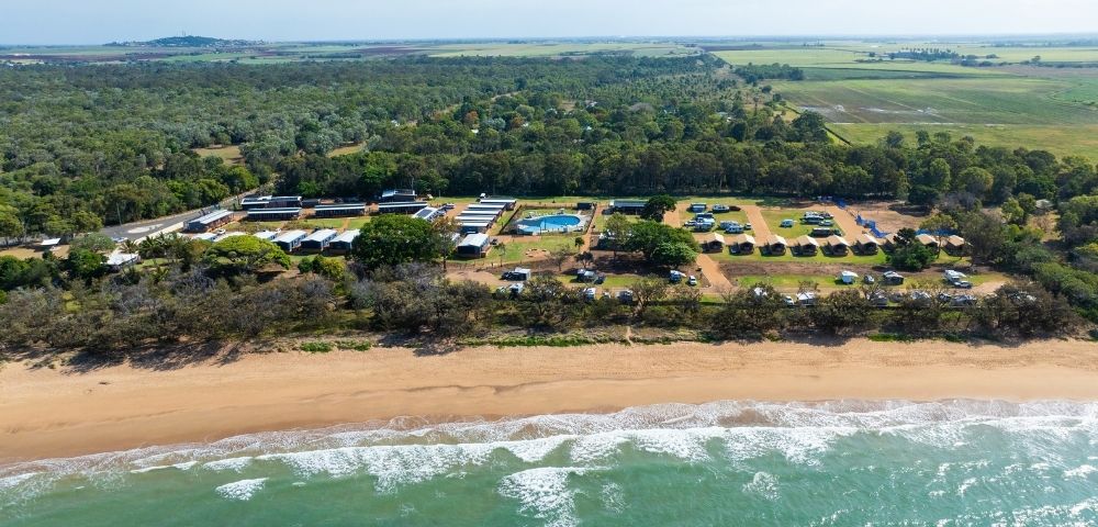Aerial view of a beachside campground with multiple cabins, a swimming pool, and scattered tents and RVs surrounded by trees and open fields in the background.