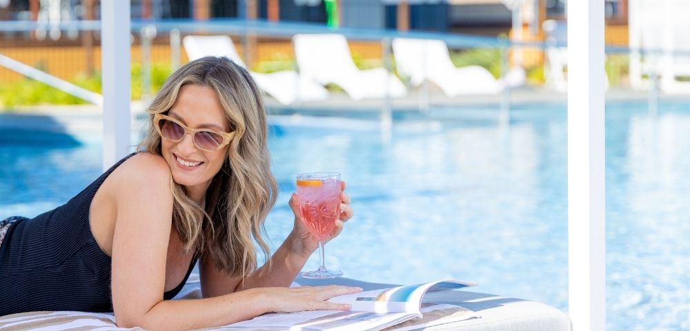 A woman wearing sunglasses and a black swimsuit lies on a chaise lounge by a pool, holding a pink drink and a magazine.