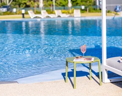 A pink cocktail sits on a green side table next to a swimming pool, with a sun hat and towel on a nearby lounge chair.