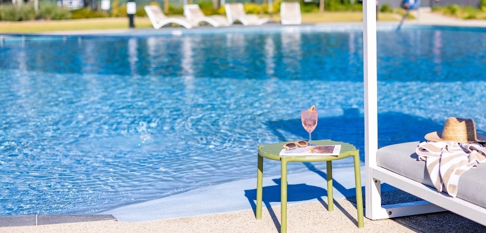 A pink cocktail sits on a green side table next to a swimming pool, with a sun hat and towel on a nearby lounge chair.