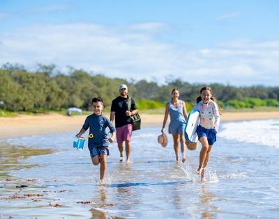 A family runs and splashes through shallow water on a sunny beach, with two children in the foreground and two adults behind them.