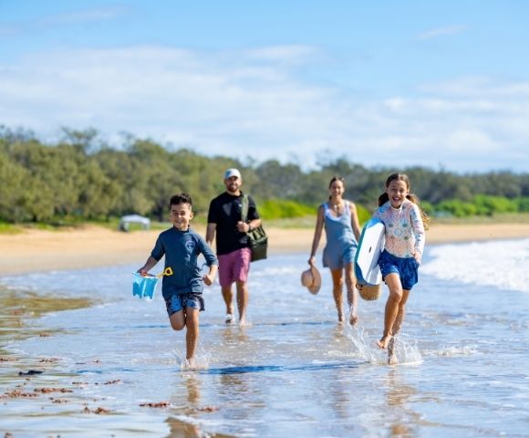 A family runs and splashes through shallow water on a sunny beach, with two children in the foreground and two adults behind them.