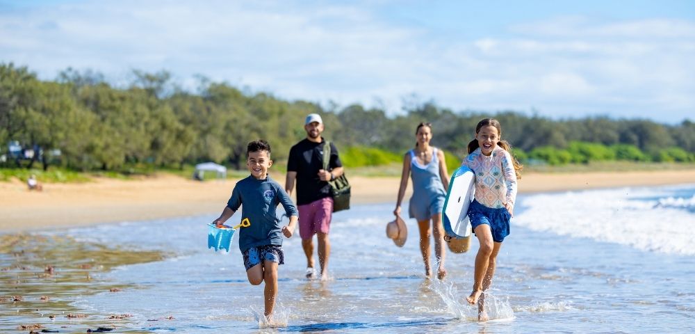 A family runs and splashes through shallow water on a sunny beach, with two children in the foreground and two adults behind them.