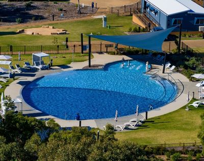 An aerial view of a large, blue, multi-level swimming pool with several people playing in the shallow end and lounging chairs around the perimeter.