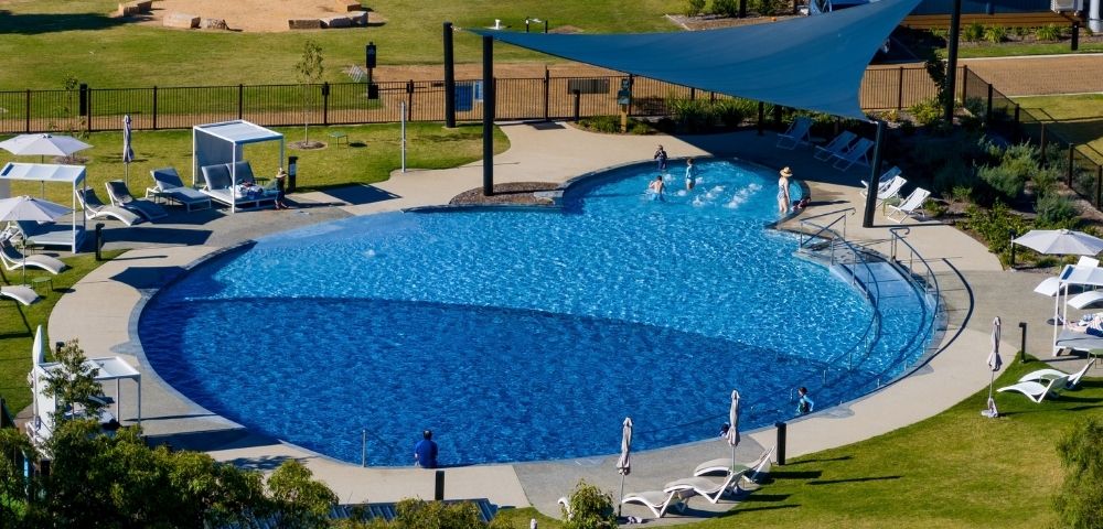 An aerial view of a large, blue, multi-level swimming pool with several people playing in the shallow end and lounging chairs around the perimeter.