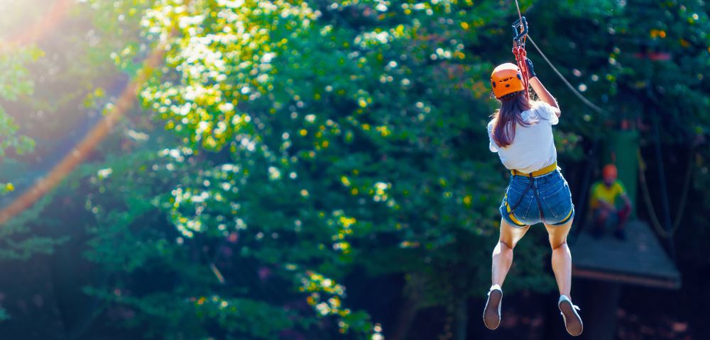A person in a white shirt and denim shorts zips through a lush green forest canopy, wearing an orange helmet and safety harness.