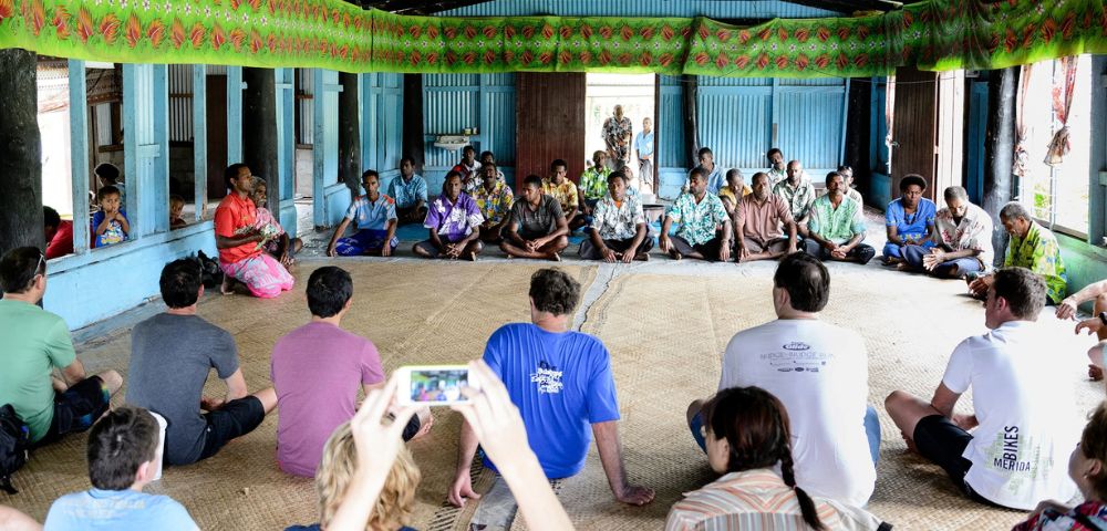 A diverse group of people, mostly men wearing casual clothing and some in traditional island attire, are seated in a circle on woven mats inside an open-sided building decorated with green and red patterned fabric.