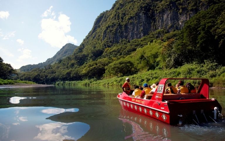 A red tour boat carrying several passengers in yellow life vests navigates a calm river, reflecting the lush green mountains and blue sky.