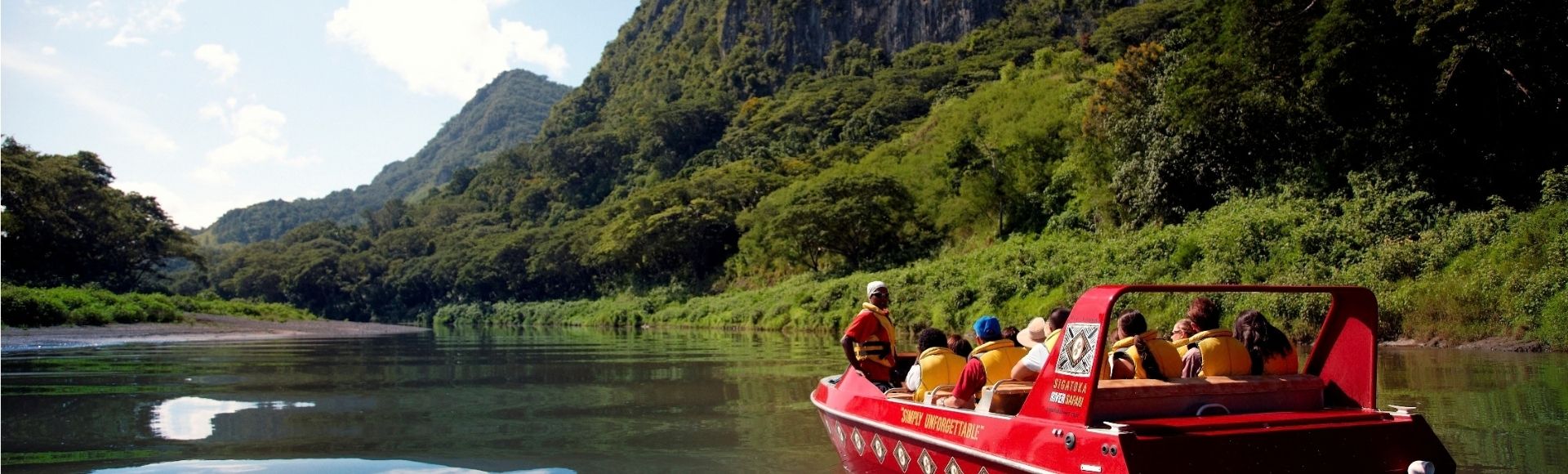 A red tour boat carrying several passengers in yellow life vests navigates a calm river, reflecting the lush green mountains and blue sky.