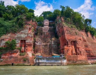 A colossal ancient stone statue of a seated Buddha carved into a red sandstone cliff face, with lush green trees on the cliff tops and a wide river in the foreground.