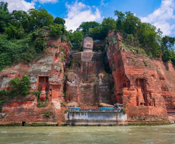 A colossal ancient stone statue of a seated Buddha carved into a red sandstone cliff face, with lush green trees on the cliff tops and a wide river in the foreground.