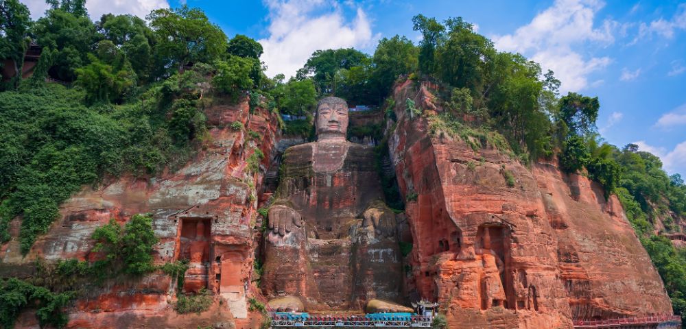 A colossal ancient stone statue of a seated Buddha carved into a red sandstone cliff face, with lush green trees on the cliff tops and a wide river in the foreground.