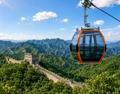 An orange and glass cable car gondola hangs suspended in the foreground against a backdrop of the Great Wall of China snaking across lush, green, mountainous terrain under a blue sky with scattered clouds.