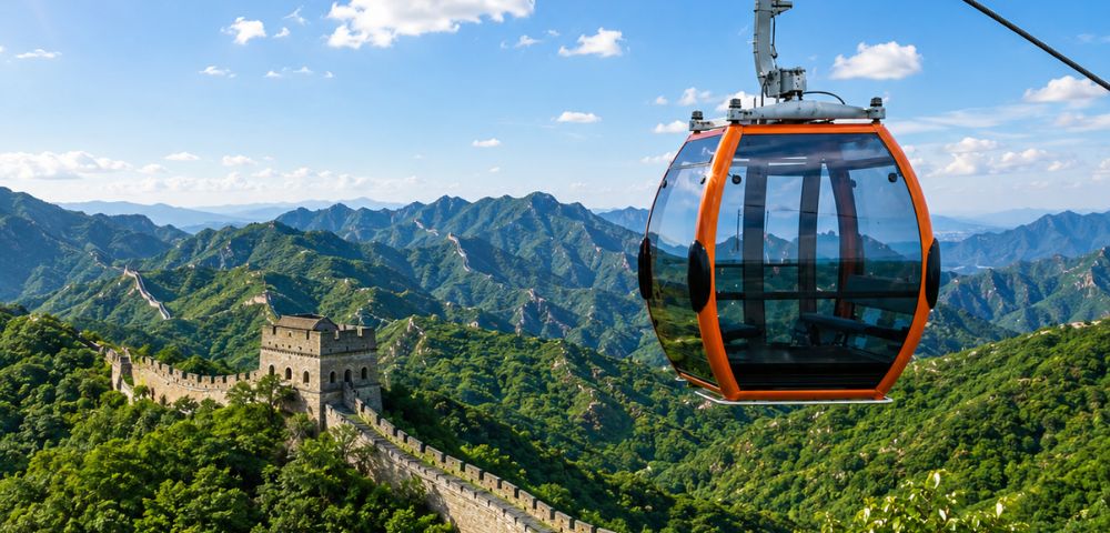 An orange and glass cable car gondola hangs suspended in the foreground against a backdrop of the Great Wall of China snaking across lush, green, mountainous terrain under a blue sky with scattered clouds.