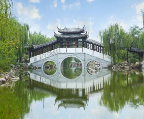 A traditional Chinese arched bridge with a covered pavilion on top, reflected in calm water surrounded by lush green willow trees.