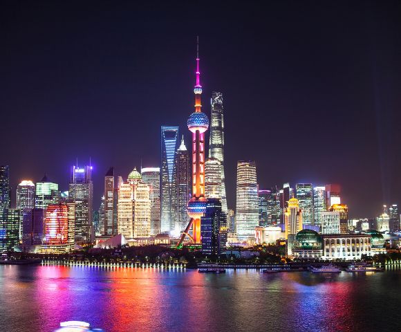 The illuminated Shanghai skyline at night with the Oriental Pearl TV Tower prominently featured, its spheres glowing red and pink, reflected in the dark water.