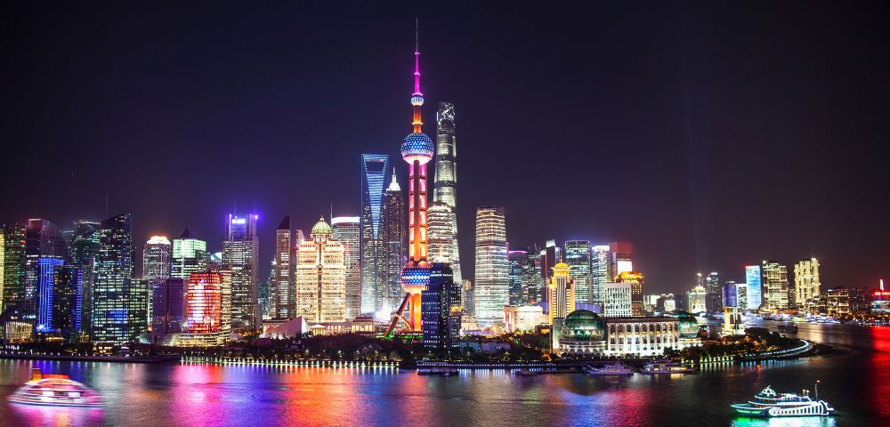 The illuminated Shanghai skyline at night with the Oriental Pearl TV Tower prominently featured, its spheres glowing red and pink, reflected in the dark water.