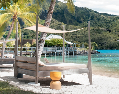 Beachside lounge chairs with canopies face turquoise water with a pier and lush, green mountains in the background.