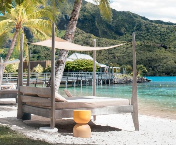 Beachside lounge chairs with canopies face turquoise water with a pier and lush, green mountains in the background.
