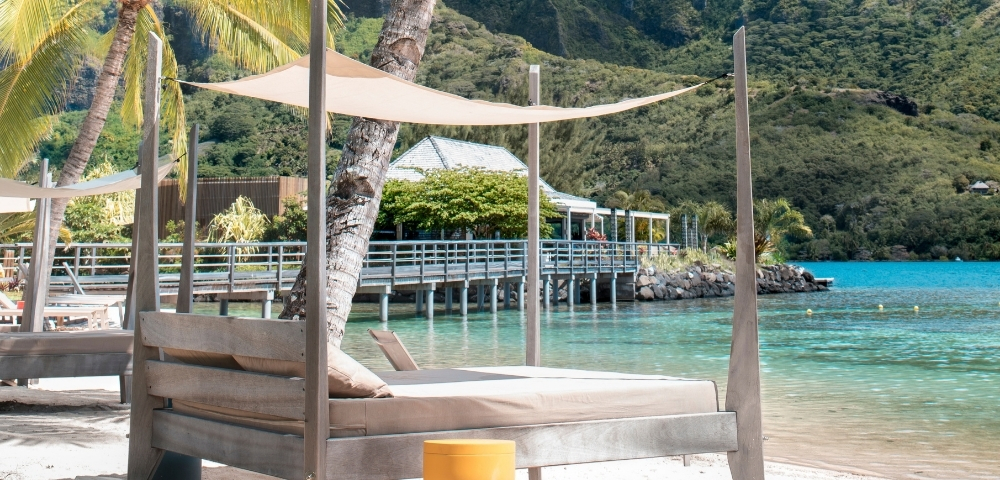 Beachside lounge chairs with canopies face turquoise water with a pier and lush, green mountains in the background.