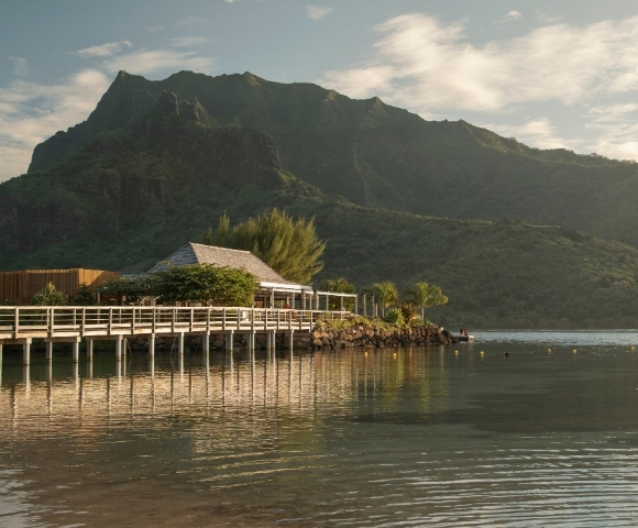 Wooden pier with a small building and lush green mountains in the background, reflected in calm water.