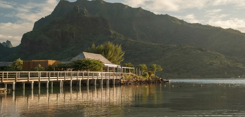 Wooden pier with a small building and lush green mountains in the background, reflected in calm water.