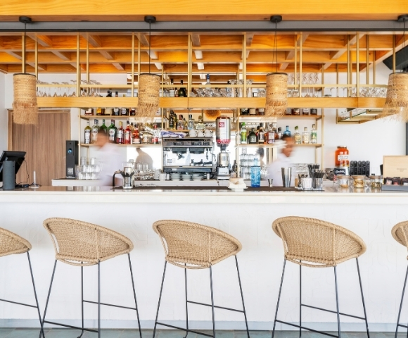 Interior of a modern bar with a long white counter, wicker stools, a professional espresso machine, and shelves stocked with bottles and glassware.