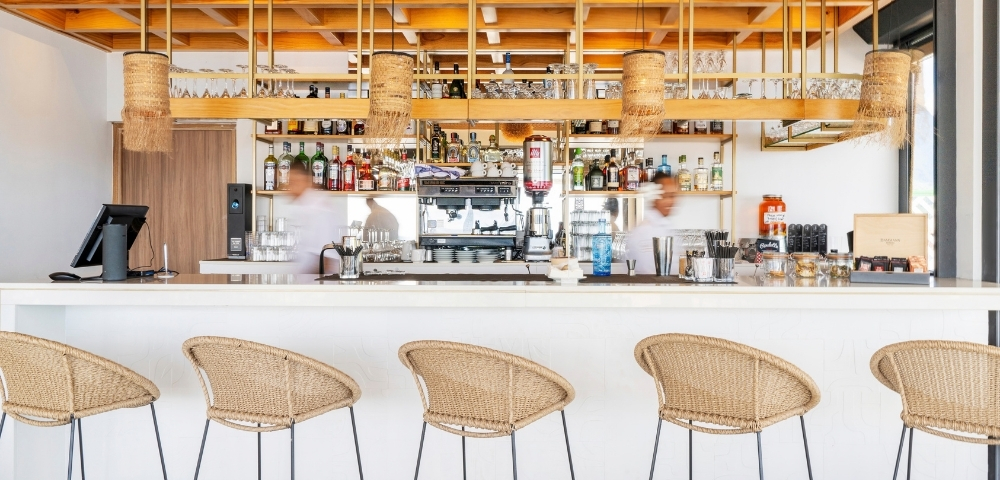 Interior of a modern bar with a long white counter, wicker stools, a professional espresso machine, and shelves stocked with bottles and glassware.