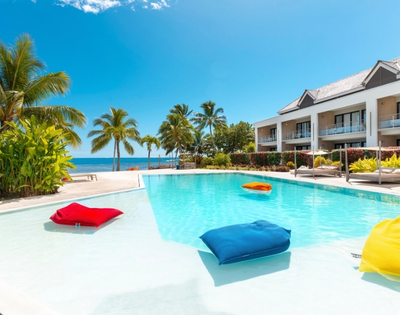 A resort swimming pool with bright blue water and white shallow edges, featuring inflatable cushions in red, blue, and orange floating in the water, with a modern hotel building and palm trees in the background under a clear blue sky.