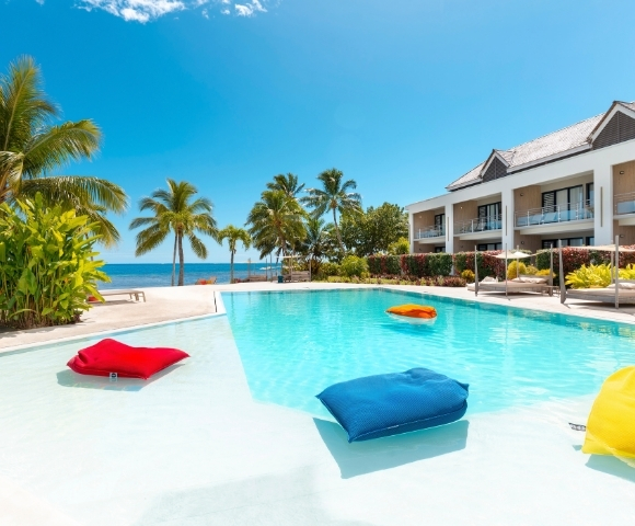 A resort swimming pool with bright blue water and white shallow edges, featuring inflatable cushions in red, blue, and orange floating in the water, with a modern hotel building and palm trees in the background under a clear blue sky.