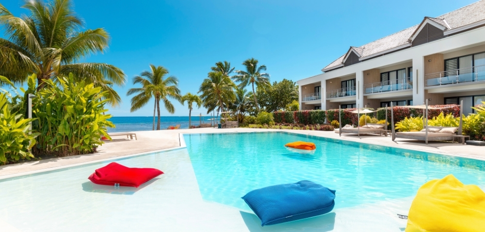 A resort swimming pool with bright blue water and white shallow edges, featuring inflatable cushions in red, blue, and orange floating in the water, with a modern hotel building and palm trees in the background under a clear blue sky.