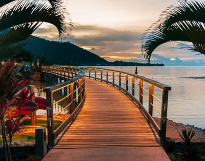 Curving wooden boardwalk with lit railings leads over calm water towards a distant, dark mountain range under a soft, cloudy sunset.