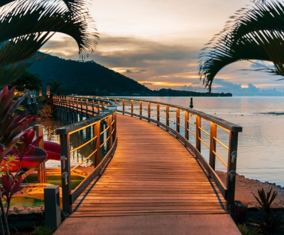 Curving wooden boardwalk with lit railings leads over calm water towards a distant, dark mountain range under a soft, cloudy sunset.