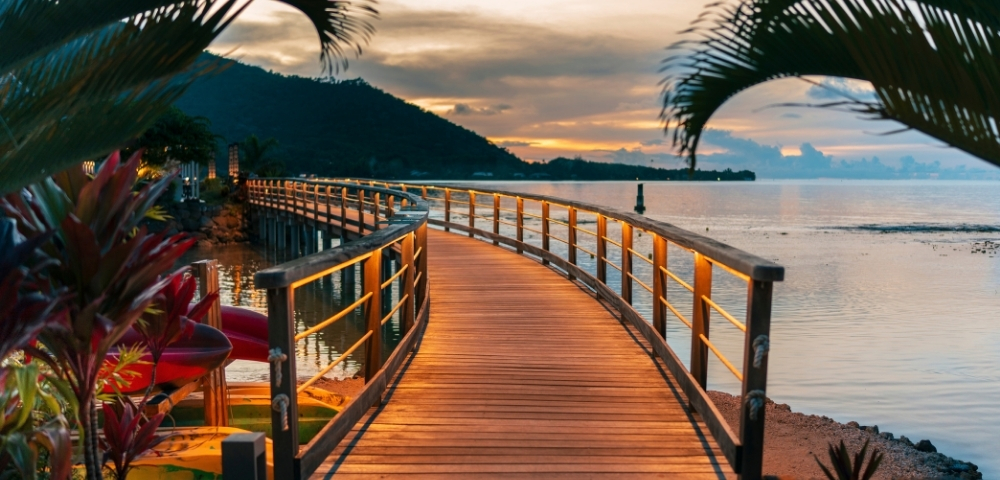 Curving wooden boardwalk with lit railings leads over calm water towards a distant, dark mountain range under a soft, cloudy sunset.