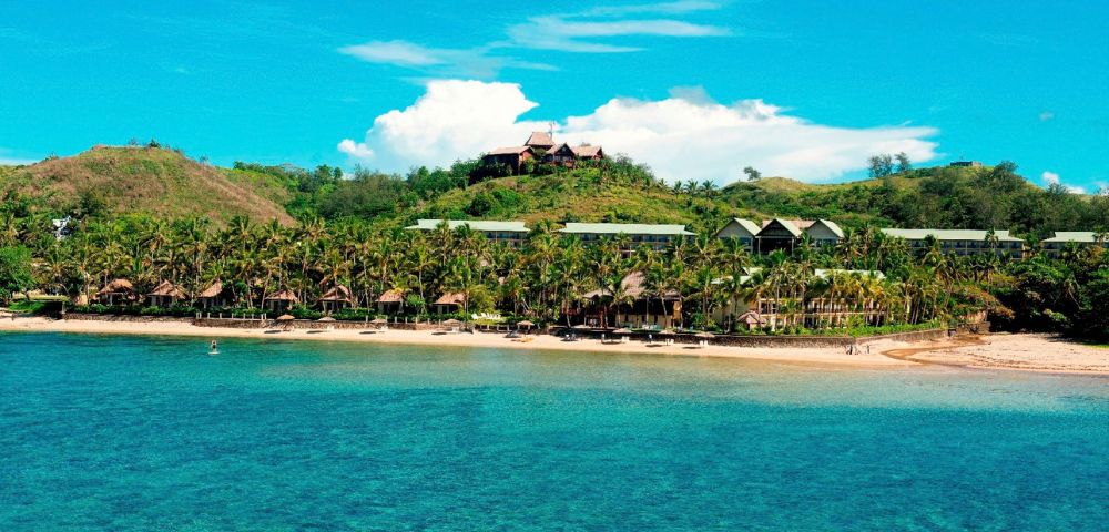 A tropical resort with bungalows and hotel buildings nestled among palm trees on a sandy beach, viewed from the turquoise ocean under a bright blue sky with puffy white clouds.